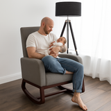 Man sitting in a chair holding a baby in a well-lit room with a lamp and window in the background.