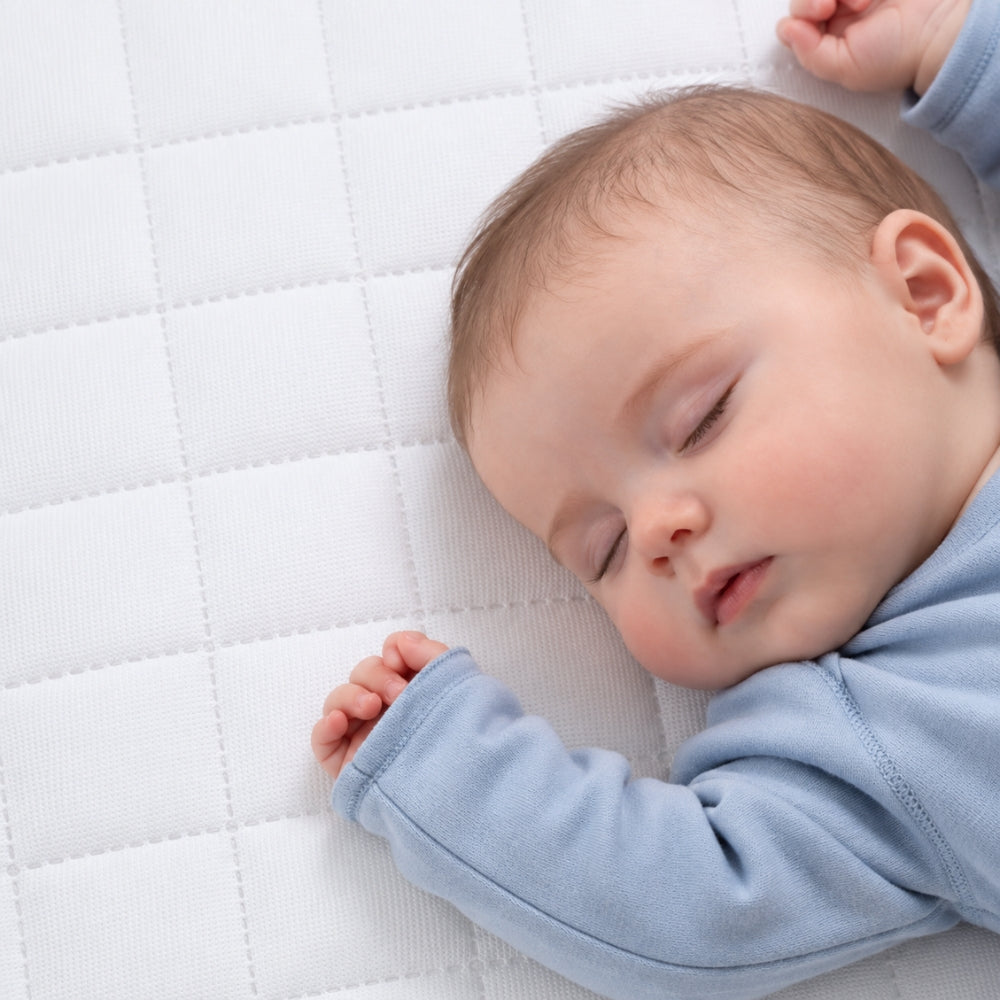 A sleeping baby wearing a light blue long-sleeve outfit, lying on a white quilted mattress or bedding surface.