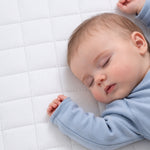 A sleeping baby wearing a light blue long-sleeve outfit, lying on a white quilted mattress or bedding surface.